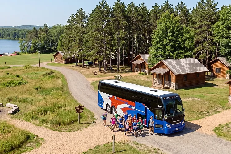 Aerial view of a Lorenz Bus Service Prevost motorcoach with campers unloading at a lakeside camp with log cabins and tall pines