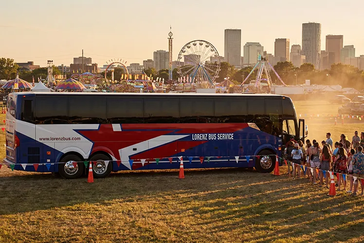 Lorenz Bus Service charter coach parked at an outdoor festival with a city skyline and Ferris wheel in the background at sunset